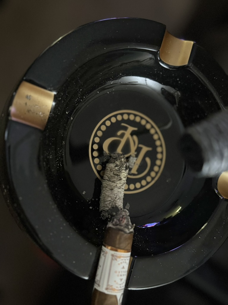 Rocky Patel LB1 Robusto with a long ash column resting across a black Rocky Patel ashtray.