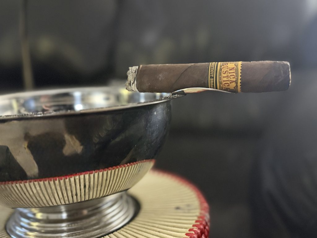 Nica Rustica Connecticut Broadleaf Toro resting on the edge of a metal ashtray with a long ash