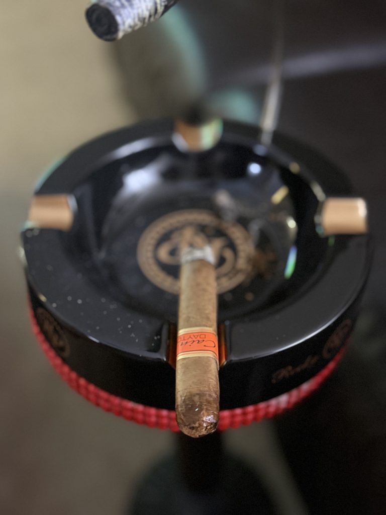 Cain Daytona Box Pressed Toro with long ash resting on edge of black Rocky Patel ashtray