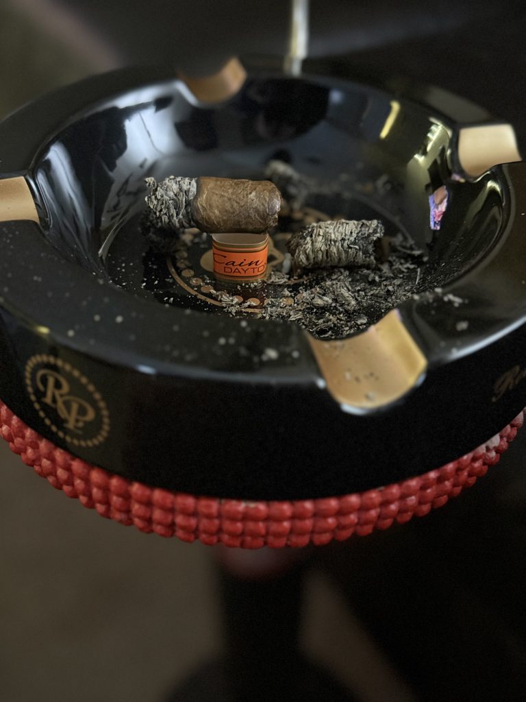Cain Daytona cigar nub on orange band in black Rocky Patel ashtray with scattered ash