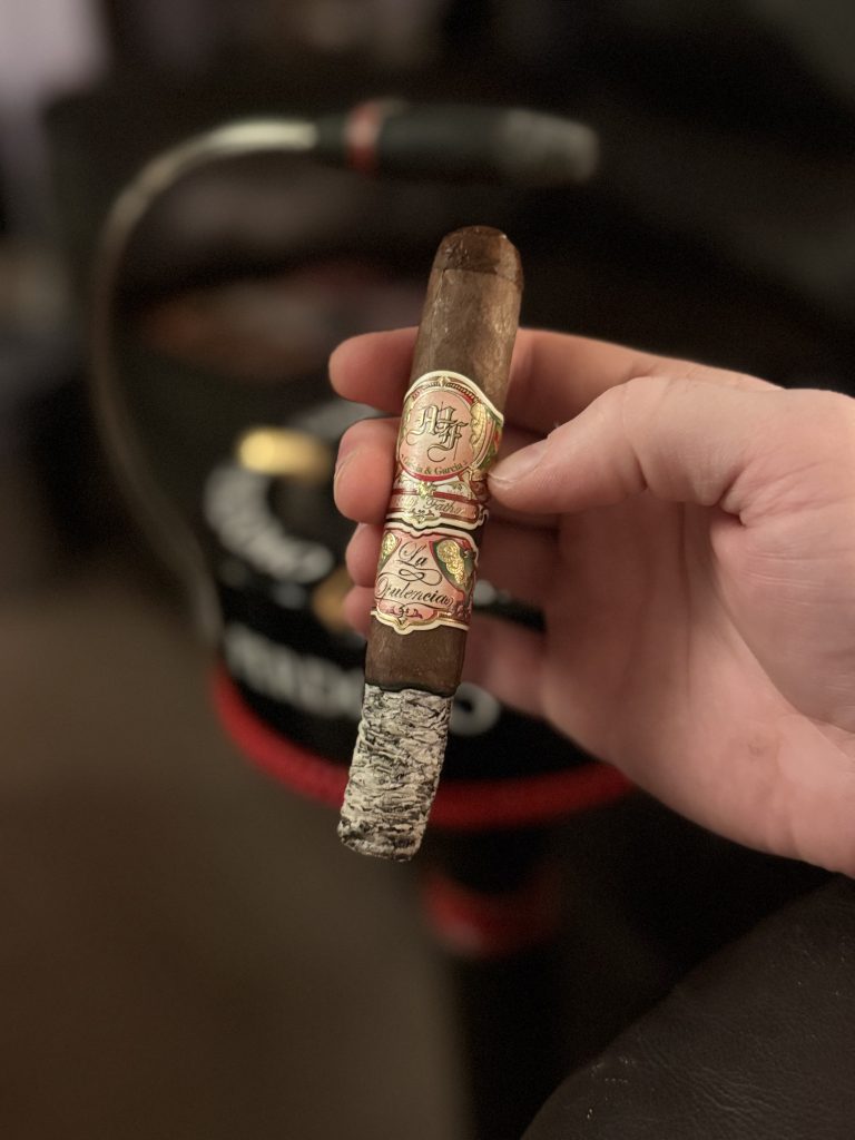 Hand holding a lit My Father La Opulencia Toro cigar with a long ash, in front of a blurred lounge background and cigar accessories.