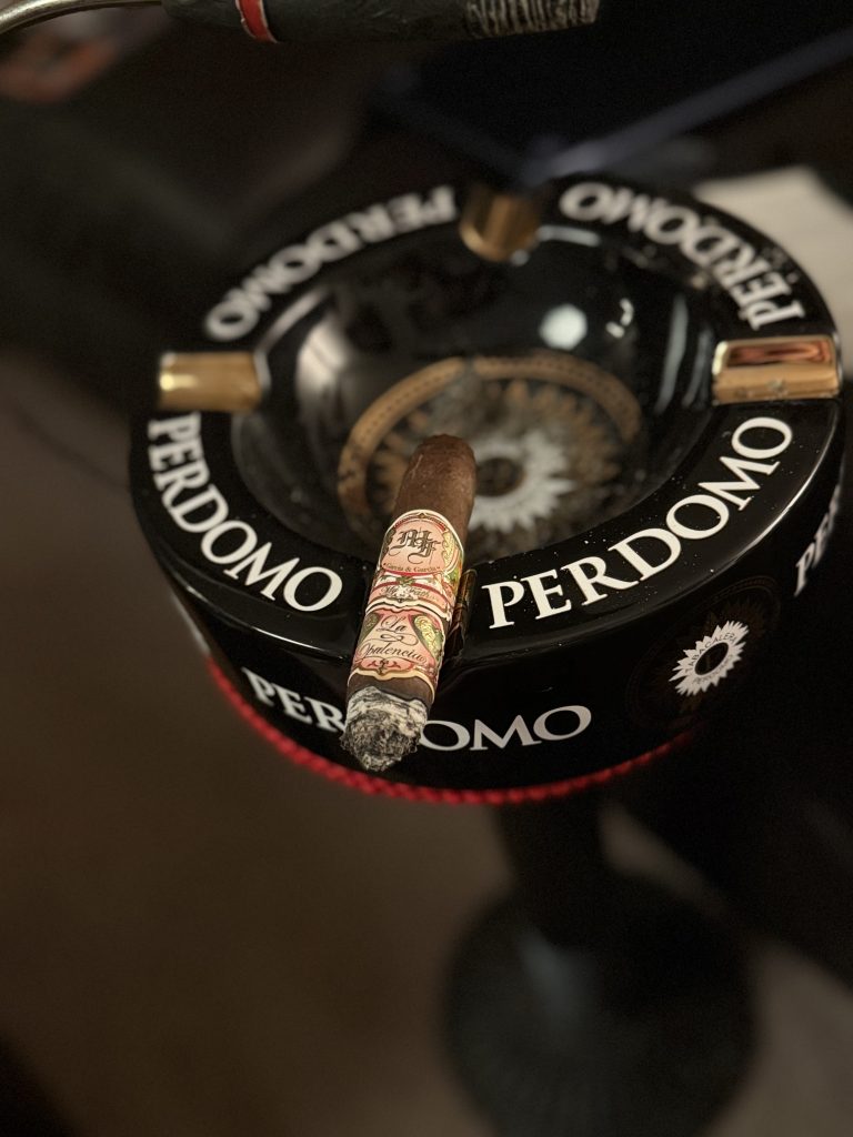 My Father La Opulencia Toro cigar resting with a long ash on a black Perdomo ashtray in a cigar lounge setting.