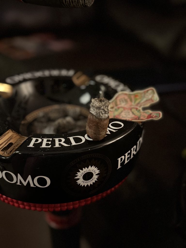 Burned nub of a My Father La Opulencia cigar resting on a black Perdomo ashtray, with the cigar band discarded beside it in a dim lounge setting.