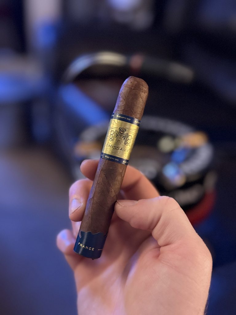Hand holding a Macanudo Emissary France cigar with gold and blue band in a dim lounge setting.