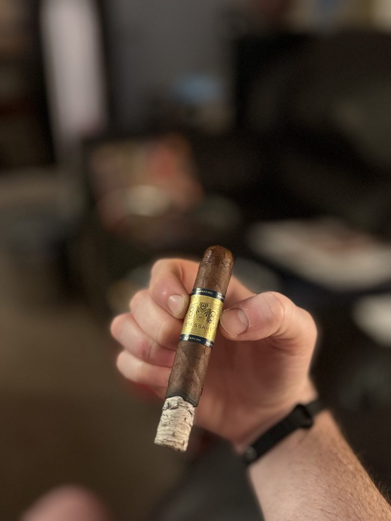 Hand holding a Macanudo Emissary France cigar with a long, firm ash in a lounge.