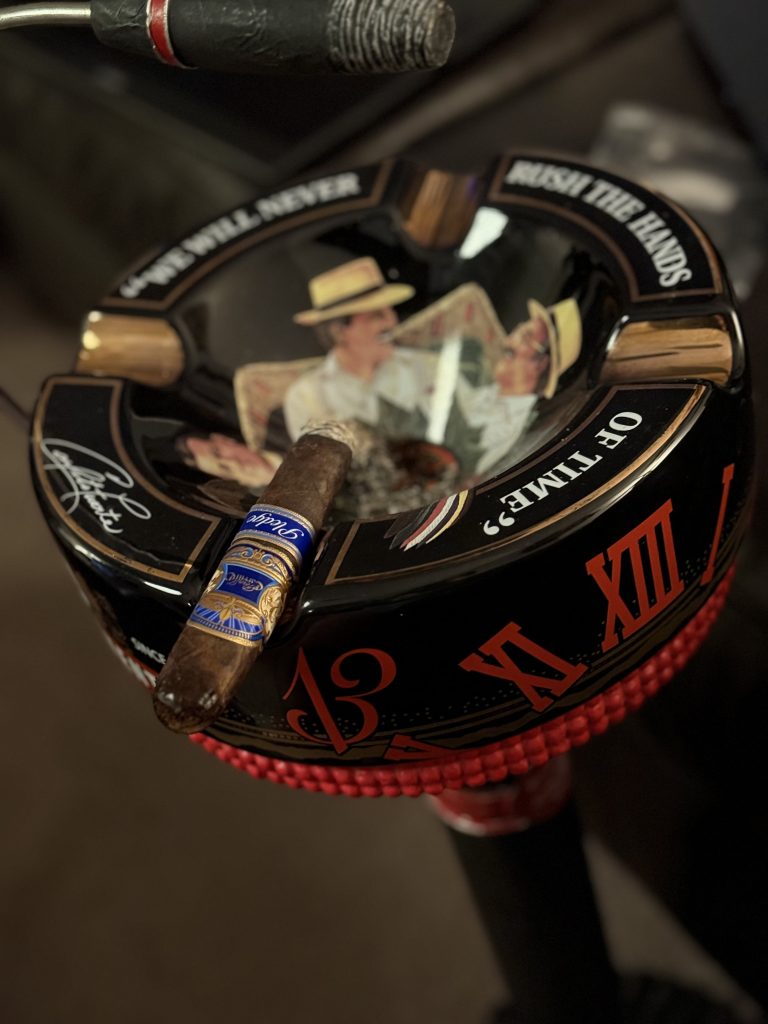 E.P. Carrillo Pledge Sojourn cigar on a black ashtray showing artwork of two men talking and the phrase “Never Rush the Hands of Time,” with the cigar’s blue band highlighted.