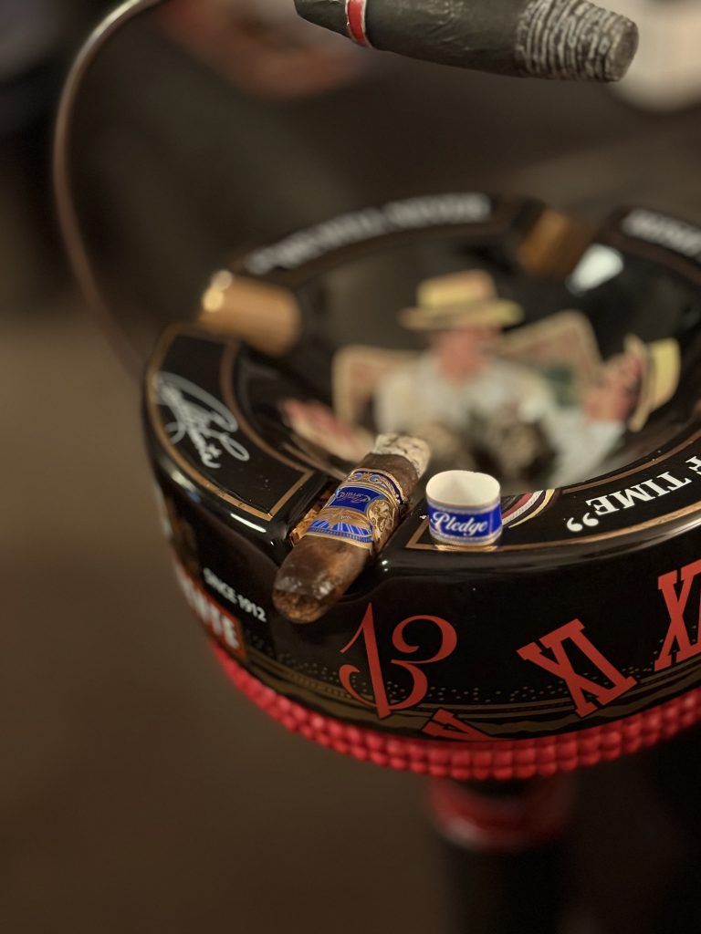 Close‑up of an E.P. Carrillo Pledge Sojourn cigar resting on a black, clock‑themed ashtray with red Roman numerals and the cigar’s blue band in focus.