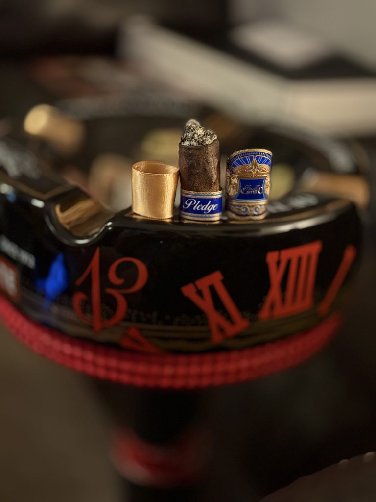 Nearly finished E.P. Carrillo Pledge Sojourn cigar nub standing upright between its blue primary band and gold foot ribbon on a black ashtray with red Roman numerals.