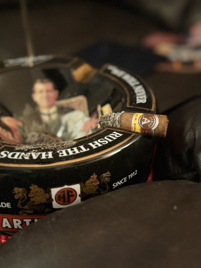 Aladino Corojo Robusto in the final third resting on a black ashtray in The Evening Draw Lounge.