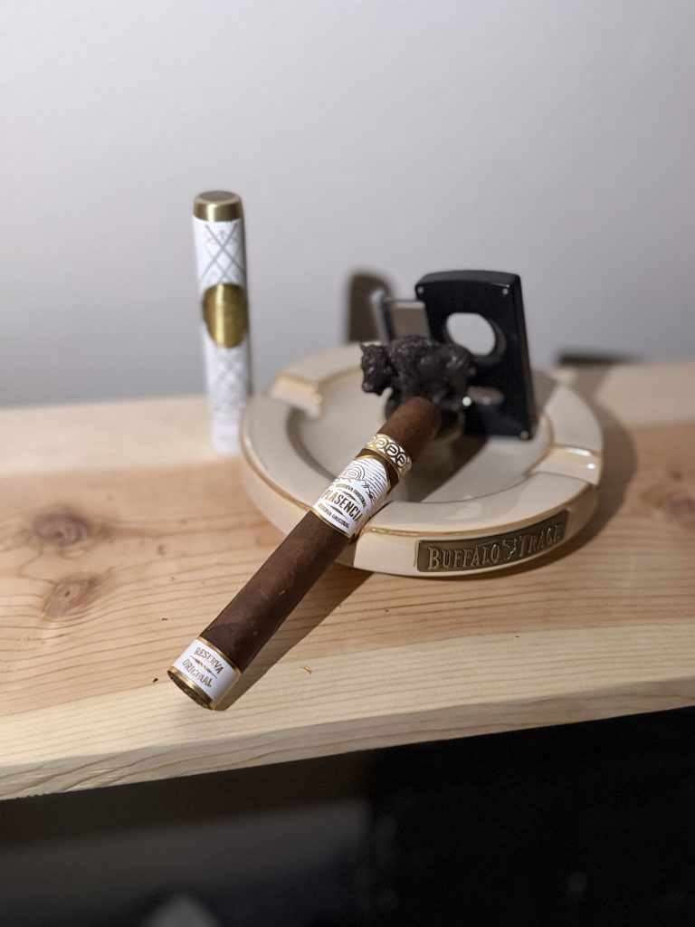 Plasencia Reserva Original Toro resting on a Buffalo Trace ceramic ashtray beside a white and gold Les Fines Lames cigar case and black double guillotine cutter before the cigar is lit.