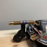 AJ Fernandez Amar Toro resting on a black cigar cutter stand before lighting, showing the purple and gold primary band and gold foot band against a clean wood tabletop.