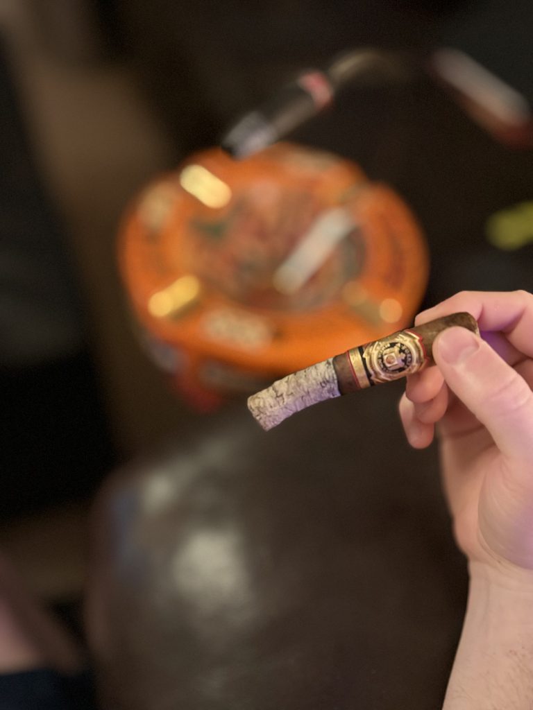Close-up of the Arturo Fuente Reserva Don Carlos Robusto with a long ash and blurred Casa Cuba ashtray in the background.