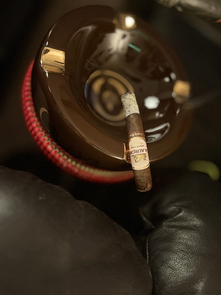 Lit La Aurora Family Creed Fuerte Sol Robusto with a long white ash resting over a brown cigar ashtray.