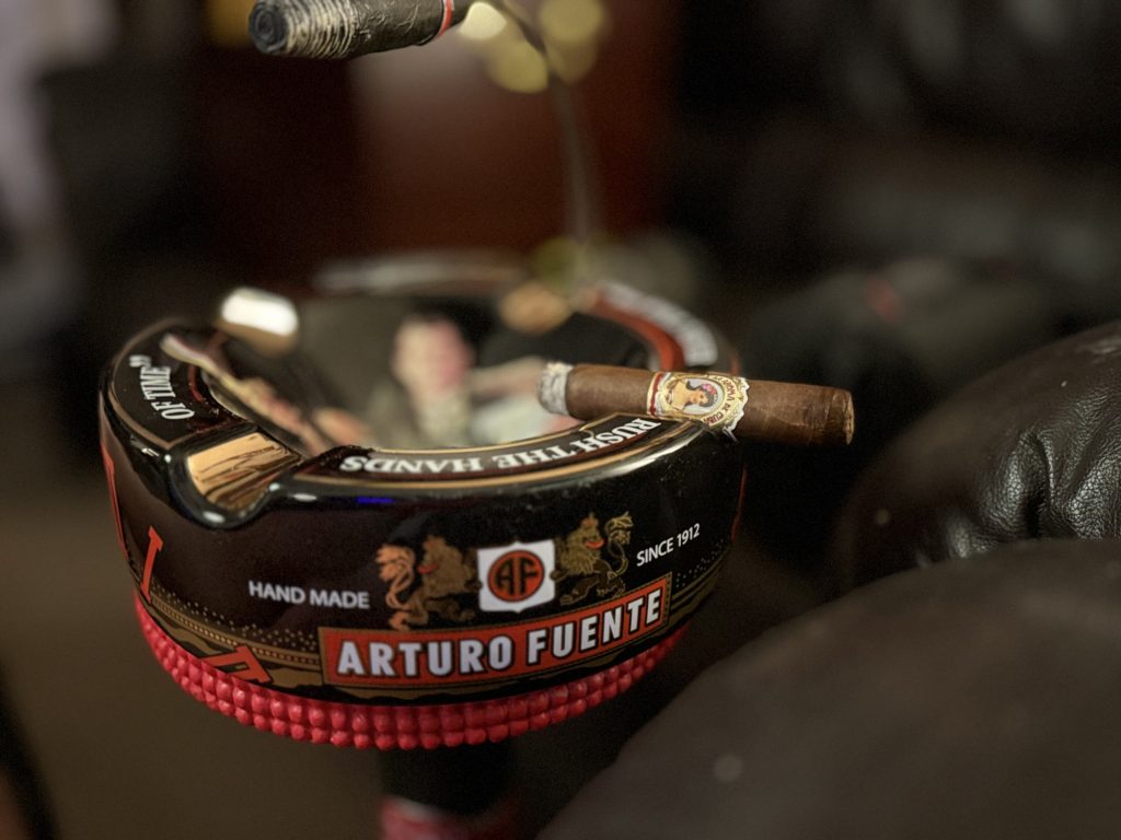 Lit La Aroma de Cuba Original Robusto resting across a black Arturo Fuente ashtray with a second smoking cigar above it.