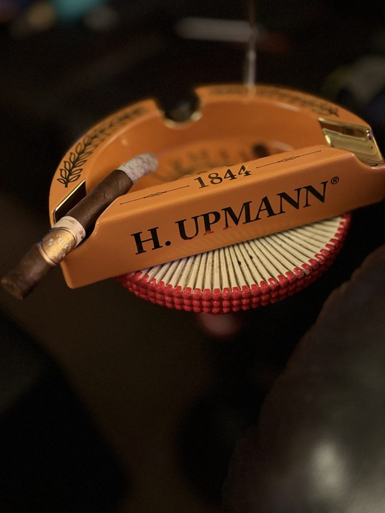 Lit H. Upmann 180 Anniversary Churchill with a long ash resting across an orange H. Upmann 1844 ashtray.