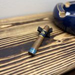 Macanudo Emissary España Robusto resting on a black cigar stand on a charred-wood table before lighting.