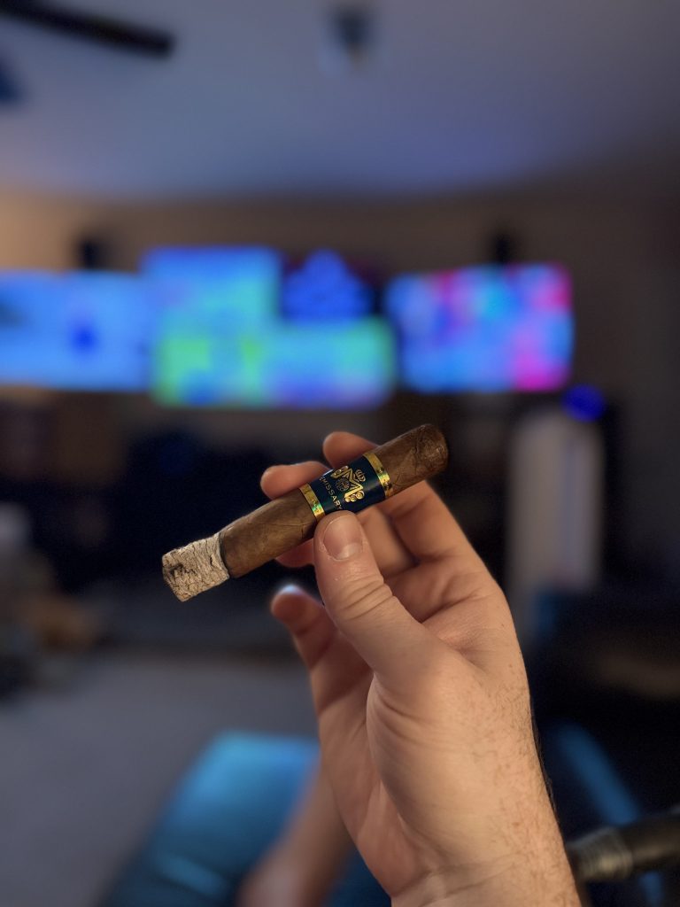 Lit Macanudo Emissary España Robusto held in hand with a long white ash and lounge televisions blurred behind it.