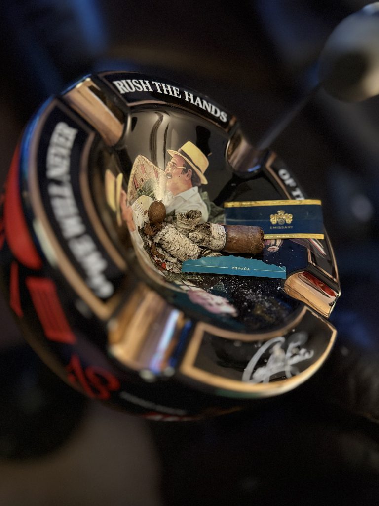 Finished Macanudo Emissary España Robusto nub and ash beside its removed navy Emissary band and turquoise España foot band.
