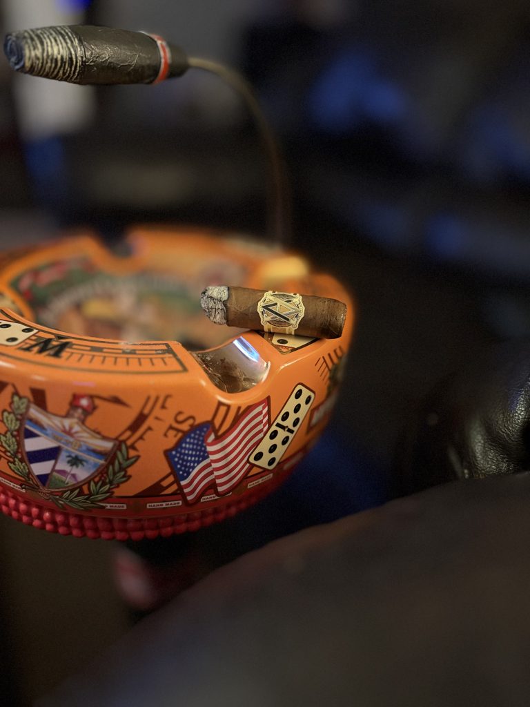 Burning AVO Classic Natural Robusto resting across an orange Cuban-themed ashtray with its ash and AVO band visible.