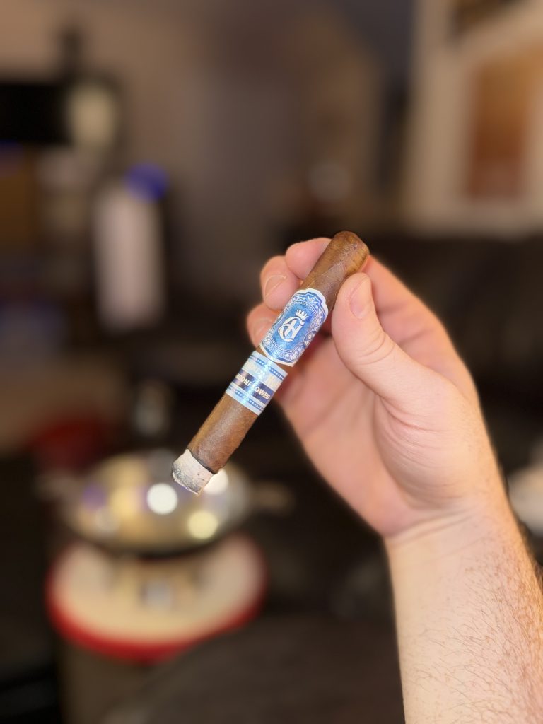 Lit Crowned Heads Moonflower Corona Gorda held by hand with an intact gray ash and blue Crowned Heads bands.