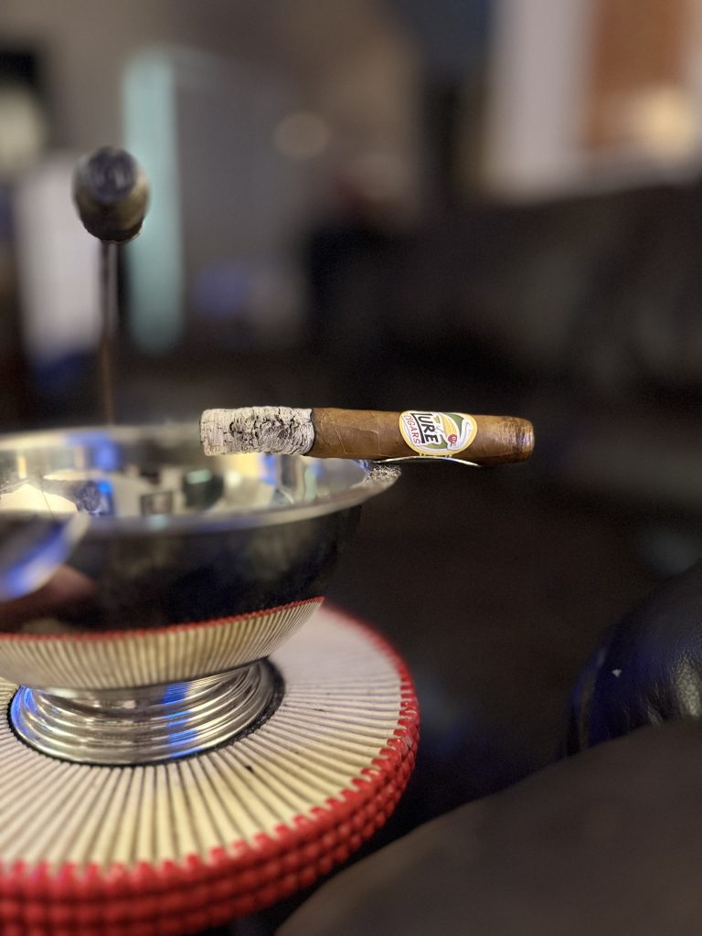 Lure Cigars Tally Allie Habano Toro resting across a metal cigar ashtray with a long ash intact.