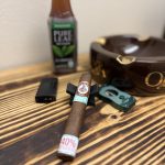 League of Fat Bastards Serie O Toro resting on a cigar stand beside a cutter, lighter, Pure Leaf unsweetened tea and Oliva ashtray.