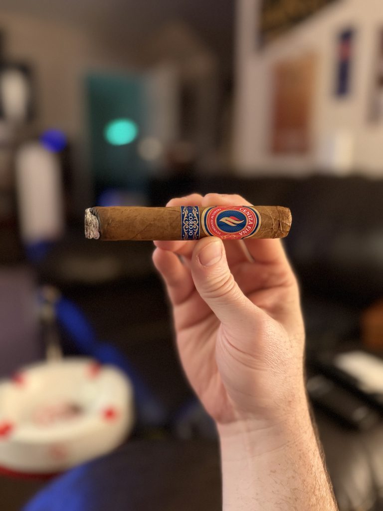 Hand holding an unlit Ceniza Fina Corojo Robusto horizontally with its blue and red band visible.