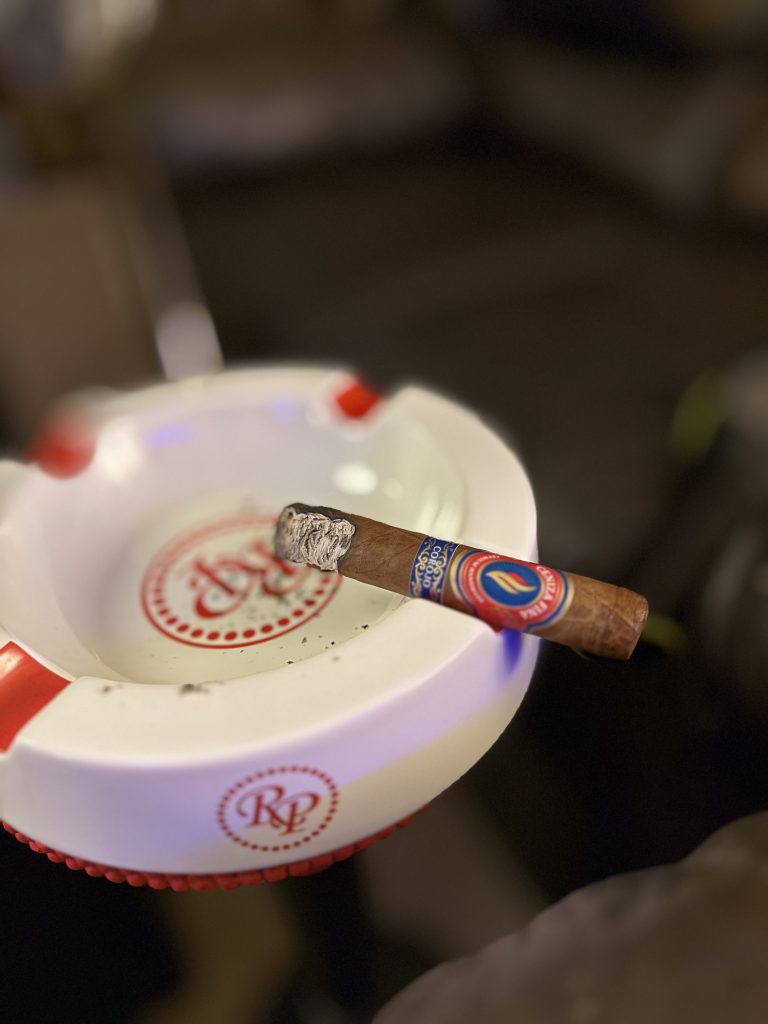 Lit Ceniza Fina Corojo Robusto resting across a white Rocky Patel ashtray with a firm gray ash.