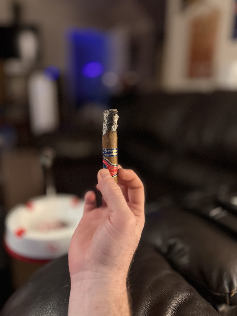Hand holding the partially smoked Ceniza Fina Corojo Robusto upright with a long ash attached.