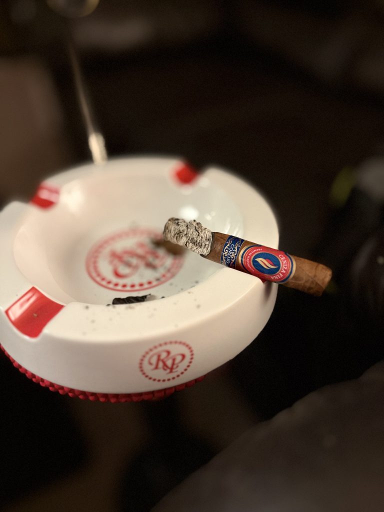 Partially smoked Ceniza Fina Corojo Robusto resting on a white ashtray with smoke rising from its ash.