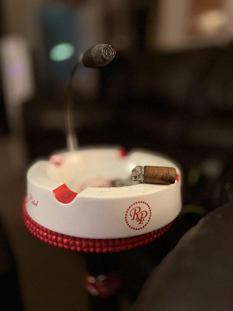 Ceniza Fina Corojo Robusto nub resting in a white ashtray beneath a drifting ring of smoke.